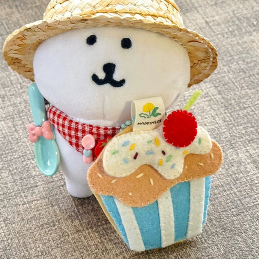 Plush toy resembling a snowman with a straw hat and cupcake, sitting on a textured surface.
