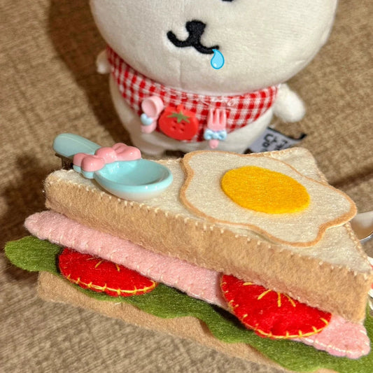 Plush toy resembling a bear with a sandwich and utensils on a textured surface