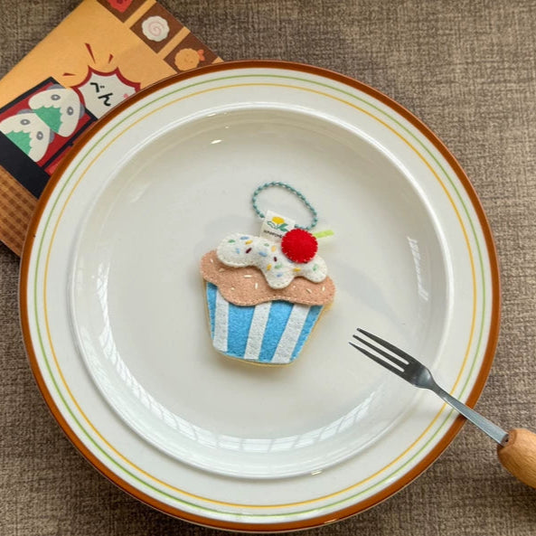 Decorative cupcake charm on a white plate with a fork, on a textured brown surface.